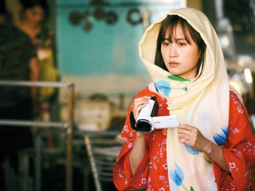 Eine weiblich gelesene Person trägt ein rot geblümtes Kleid und einen hellen Schal um den Kopf gewickelt. In der Hand hält sie eine weiße Videokamera.